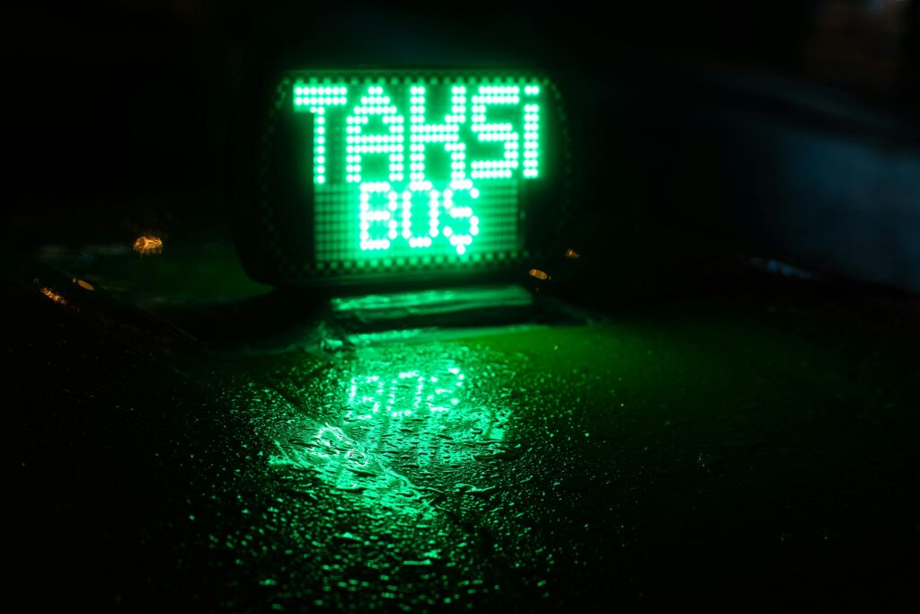 Close-up of a bright green neon taxi sign reflecting on a wet car roof at night.
